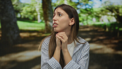 Fototapeta premium Woman clasps hands under chin and gazes anxiously to the side beneath sunlit green canopy in forest; concern.