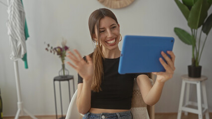 Hispanic woman smiles as she holds a blue tablet and waves hand inside a modern building; connection joy.