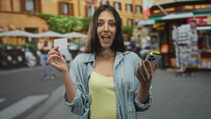 Woman holding creditcard and smartphone on street in a city market, showing card with hand and smiling; mobile payment convenience happy.