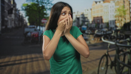 Fototapeta premium Woman hands covering mouth gesture on city street by parked bicycles and canal, wearing green shirt; shock vulnerability.