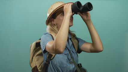 Young woman wearing safari hat and backpack holding binoculars to eyes in turquoise studio  curiosity adventure. © Krakenimages.com