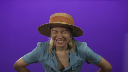 Woman in denim shirt wearing straw hat squints eyes and leans forward in pain in purple studio; distress.