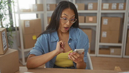 Woman fist pump while checking smartphone amid stacked shipping boxes in building packing area; small business joy.