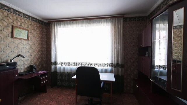 cozy room with patterned wallpaper, a large window with sheer curtains, an armchair, a desk with a lamp and printer, a cabinet, and a simple table with a chair. Warm, homely atmosphere.