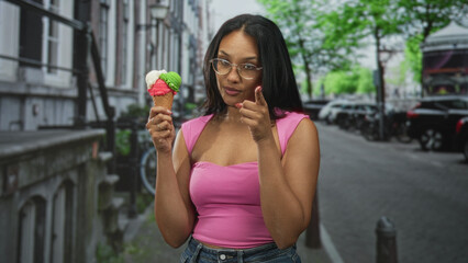 Young hispanic woman holding a three-scoop ice cream cone, wearing pink top and glasses, touches her chin and points finger while standing on a city street; playful summer fun.