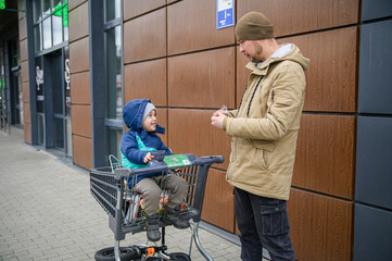 Father and son spending time together during a winter grocery trip, walking with a shopping cart, sharing snacks, and enjoying everyday family moments outdoors near a store.