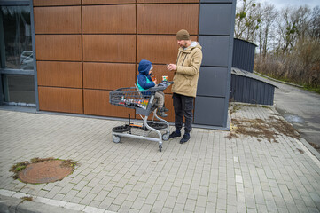 Father and son spending time together during a winter grocery trip, walking with a shopping cart, sharing snacks, and enjoying everyday family moments outdoors near a store.