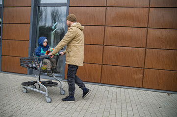 Father and son spending time together during a winter grocery trip, walking with a shopping cart, sharing snacks, and enjoying everyday family moments outdoors near a store.