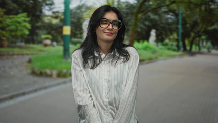 Fototapeta premium Young hispanic woman wearing glasses tilts head with a slight smile on urban street; quiet contemplation.