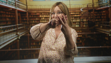 Middle age woman forms hashtag with fingers inside library building wearing glasses and a floral blouse; playful learning.