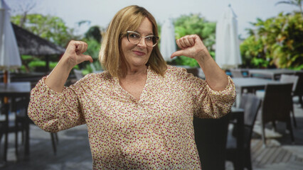 Senior woman with eyeglasses showing thumbs pointing to chest in outdoor restaurant building patio wearing a patterned blouse and glasses; personal pride.