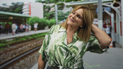 Fototapeta premium Middle age woman with blonde hair and glasses smiling, hand to neck and tilting head on a train platform beside tracks at station building, wearing green leaf blouse; confidence travel leisure.