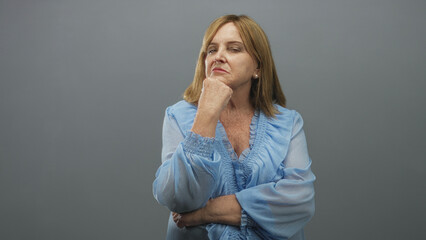Woman resting chin on fist and folding arms, wearing light blue ruffled blouse and pearl earring in studio with gray backdrop; confidence poise contemplation.