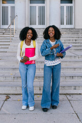 Fototapeta premium Two happy female students holding books and smiling while walking down stairs on a university campus