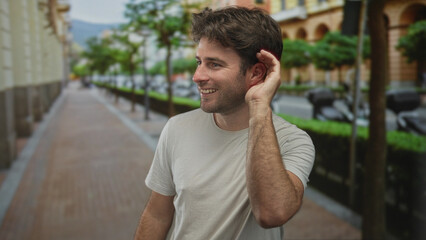 Young hispanic man smiling cupping ear to listen while standing on city street near trees and...