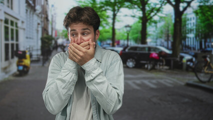 Man with hands covering mouth on a city street, glancing sideways and holding a muffled expression; shock silence.