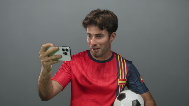 Man holding soccer ball with hand while taking a selfie with smartphone in gray studio; team spirit excitement. - Powered by Adobe