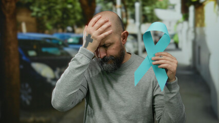 Bald man with beard holding blue cancer ribbon while standing in an urban street, expressing concern and thoughtfulness, symbolizing awareness and solidarity.
