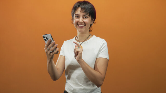 Woman points finger to smartphone in studio, smiling and tapping screen while holding phone with left hand and wearing orange pendant; joyful connection. - Powered by Adobe