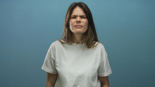 Woman frowning in discomfort over a plain blue background wearing a white t-shirt suggesting surprise or confusion in a close-up portrait setting with isolated blue.