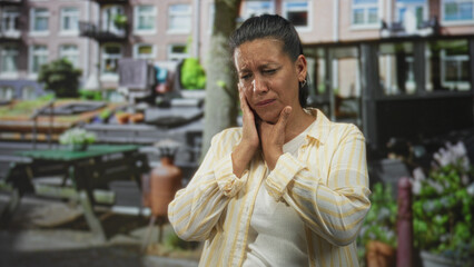 Fototapeta premium Woman holding jaw with both hands, eyes closed and grimacing in pain on a residential street with picnic table and houseboat visible; toothache pain.