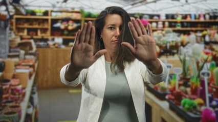 Obraz premium Woman holds palms forward in a stop gesture inside a market building filled with pottery shelves and colorful planters, wearing white jacket and knit top; defiance boundary.