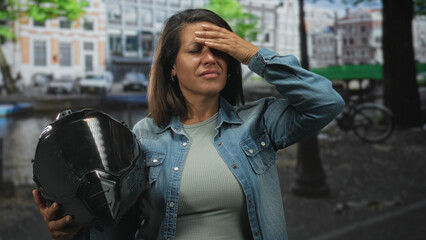Fototapeta premium Middle age hispanic woman holding glossy black motorbike helmet, hand pressed to forehead for headache beside canal street with parked bicycles; exhaustion.
