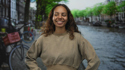 Woman smiling with hands on hips on street by amsterdam canal wearing casual sweater, posing for camera; urban confidence travel.