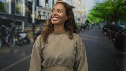 Fototapeta premium Young african american woman smiling with teeth visible on leafy city street, wearing casual sweatshirt and facing camera with relaxed posture; simple joy.