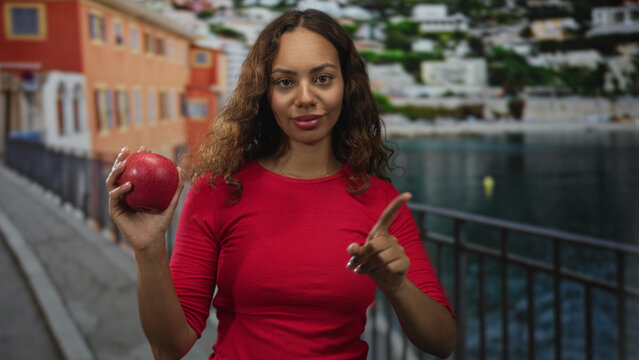 Woman holding red apple and points finger upward on a seaside street promenade near railing and waterfront, wearing red top; healthy choice confidence.