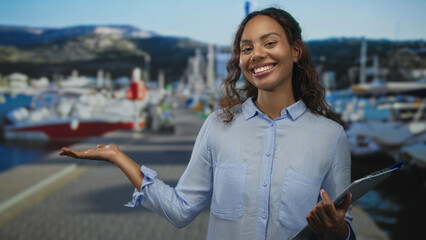 Woman smiling with palm up holding a clipboard on a marina street beside boats and dock walkway;...