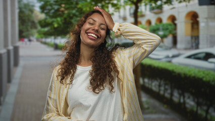 Obraz premium Woman smiling with hand on head and waving while standing on a busy city street lined with trees and parked cars; carefree joy confidence.