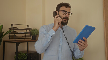 Hispanic man in office using telephone and tablet while working indoors with plants and documents in background, conveying business and communication theme.