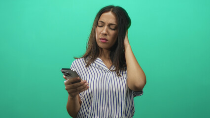 Hispanic young brunette woman holding smartphone, hand to hair and looking at screen in teal studio; concern distraction.