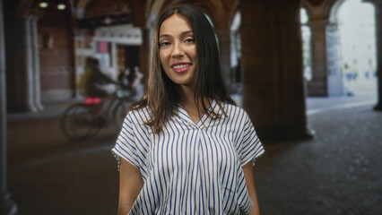 Fototapeta premium Hispanic young brunette woman smiling with face and shoulders visible in a striped shirt beneath stone arcade arches on street; confidence ease.
