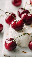 Glossy red cherries on soft goat cheese wheel over cold white marble surface with blurred fruits in background, concept of elegant gourmet summer dessert styling