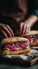 Close-up of vietnamese banh mi sandwich with pickled red onions and herbs on rustic board while asian male chef assembles food in dark blurred kitchen, concept of authentic street food culture