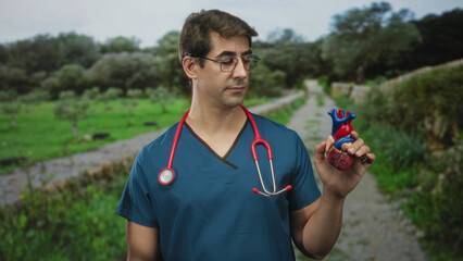 Naklejka premium Man doctor in scrubs holds anatomical heart model in right hand, stethoscope on neck, standing on forest path; medical education serious.