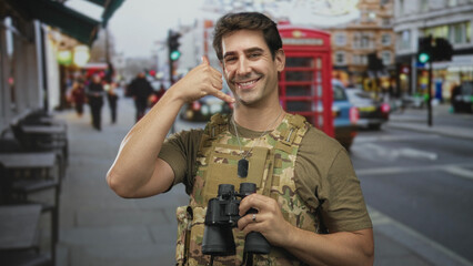 Man soldier in camouflage vest holding binoculars and making call hand gesture on a street in...