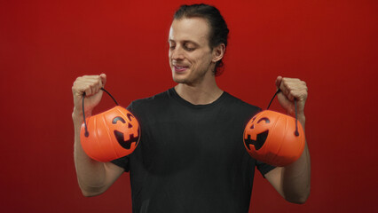 Young hispanic man with long hair holding two smiling jack o lantern pumpkin buckets by hands in a red studio; playful halloween fun.
