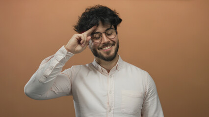 Obraz premium Young hispanic man with beard and glasses in white shirt posing against an orange background, smiling and gesturing with fingers on head, creating a positive and friendly vibe.