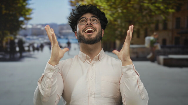 Young man with beard celebrating outdoors on a sunny street, wearing glasses and a white shirt, exuding happiness and success.