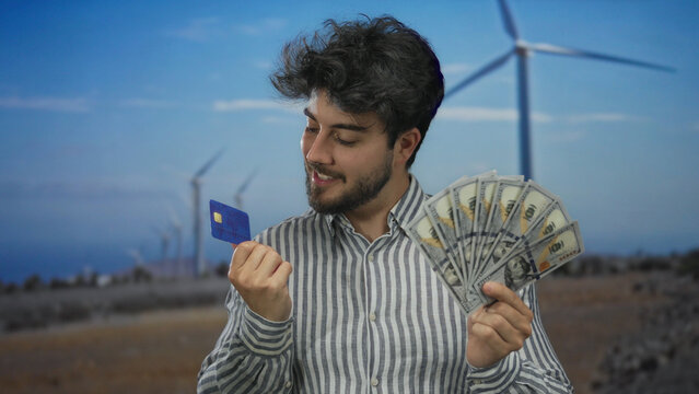 Young man with beard holding us dollars and credit card stands smiling in a field with windmills under blue sky, illustrating finance and renewable energy themes.