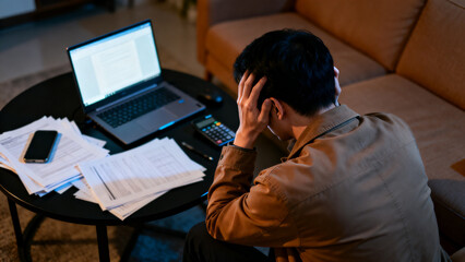 Man sitting on sofa holding his head in stress surrounded by laptop documents and calculator on cluttered table