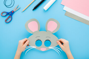 Little child girl hands holding gray mouse face mask. Scissors, marker, application paper, elastic string on light blue table background. Pastel color. Closeup. Point of view shot. Top down view.