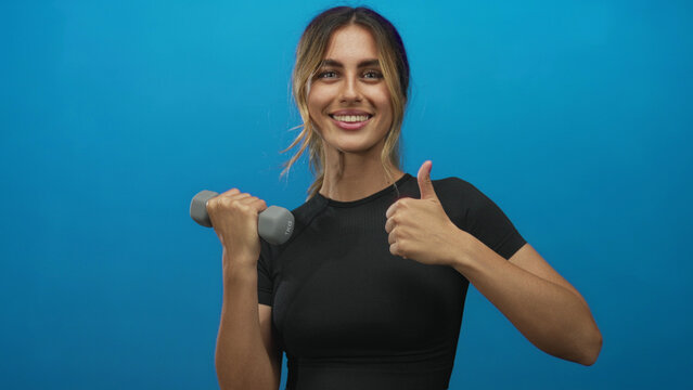 Blonde woman holds dumbbell in right hand and shows thumbs up in studio; fitness confidence motivation energy.