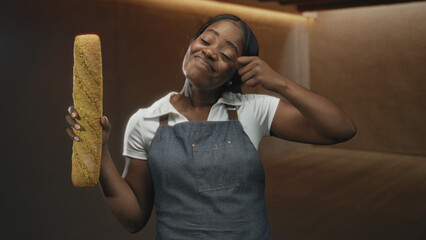 Woman baker holding a baguette while touching her temple, smiling with eyes closed in a warm bakery space wearing a denim apron; baking joy.