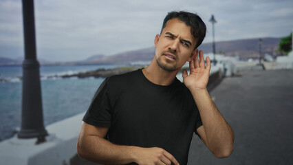 Young man standing on a seaside promenade, listening intently with focus against a backdrop of sea and distant hills, embodying curiosity and contemplation of nature's sounds.