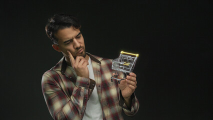 Young hispanic man in plaid shirt holding small shopping cart thinking over isolated black background conceptually illustrating online shopping or business decisions