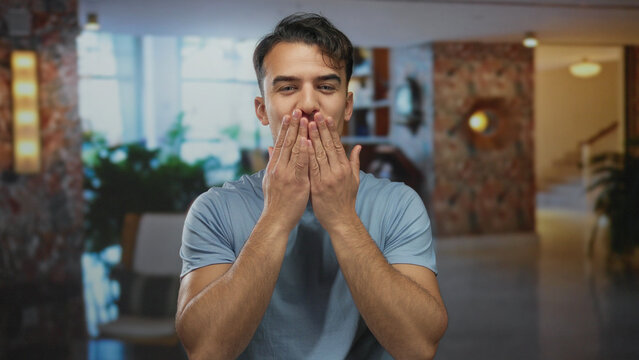 Young hispanic man in blue shirt blowing kiss to camera in modern hotel interior with plants and bright atmosphere suggesting cheerful mood and welcoming gestures.